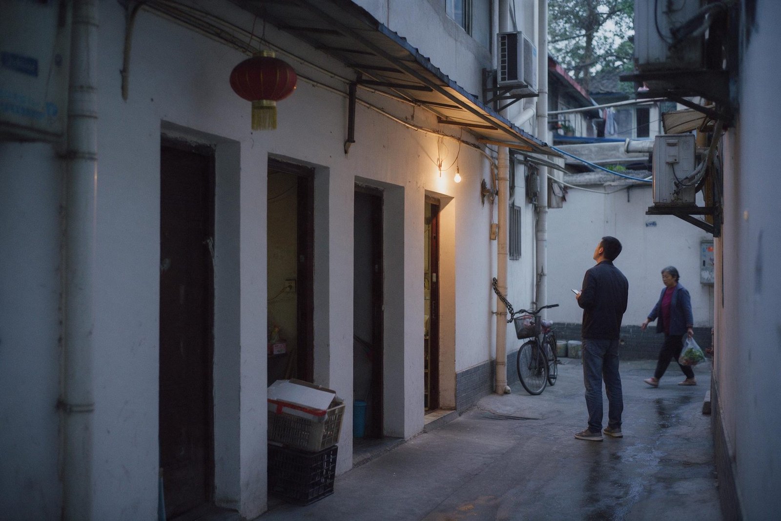 A man stands in a narrow Chengdu lane at dusk looking at four plain doorways, one lit by a single bulb.