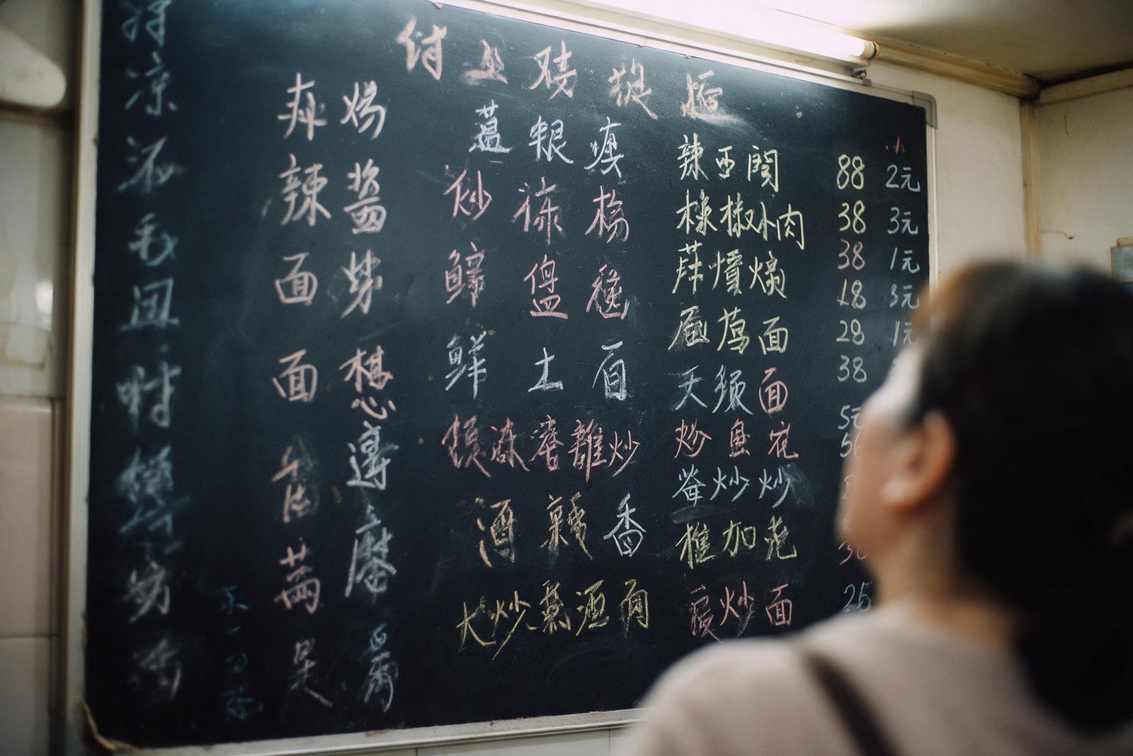 A handwritten Chinese menu board on a restaurant wall, a customer looking up at it in the foreground.