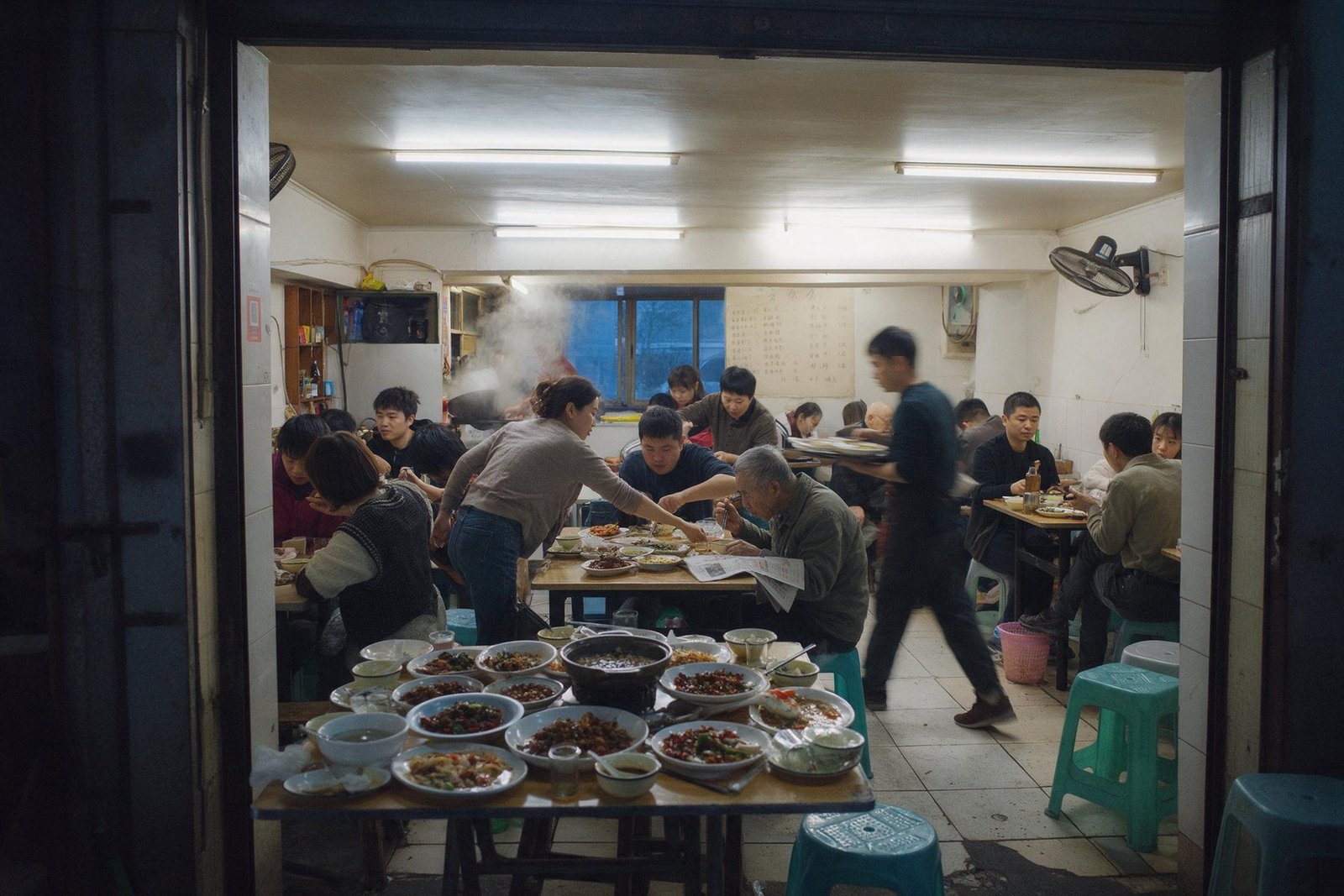 A busy small restaurant at dinner, dishes crowded on the nearest table and a waiter crossing with a tray.
