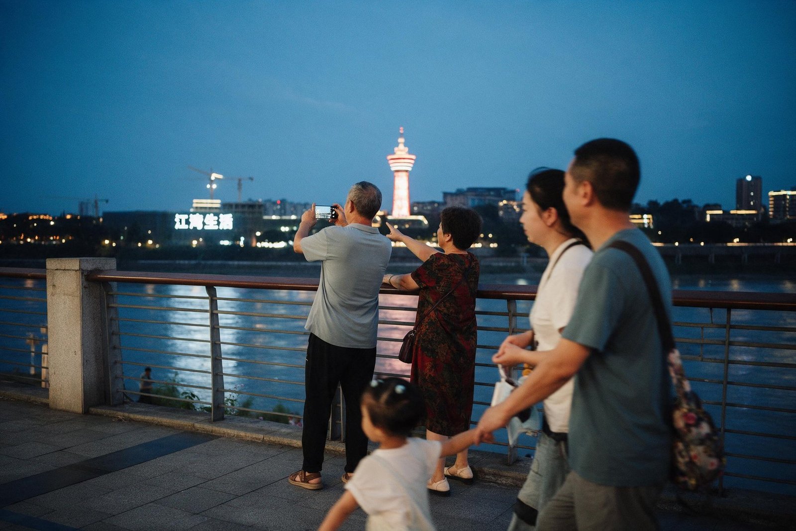 A couple photograph the far bank from a riverside promenade at blue hour as a family walks past.