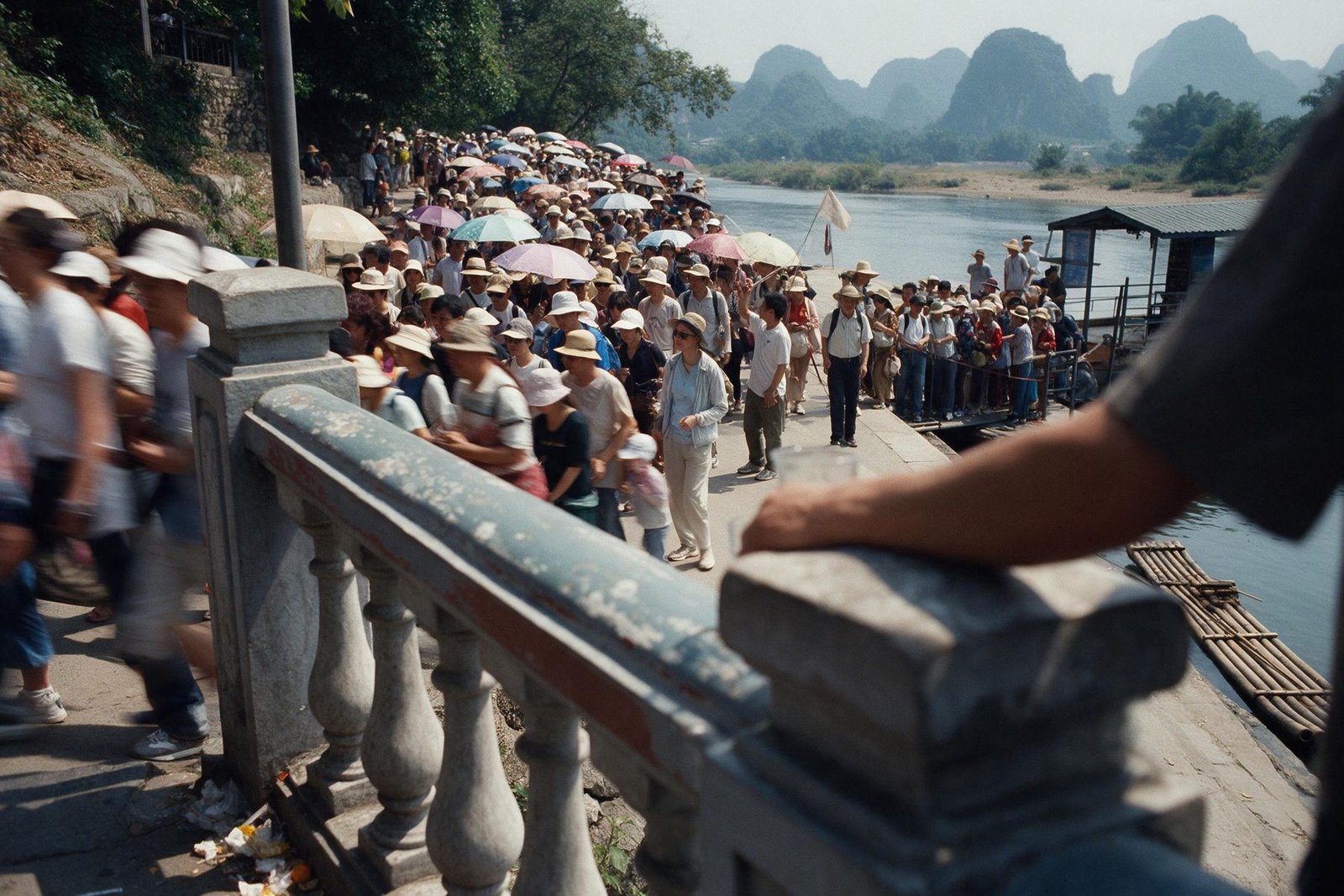 A crowded riverside path below limestone hills seen from above on a holiday morning, with sun umbrellas, a guide's flag and a queue at a raft jetty.